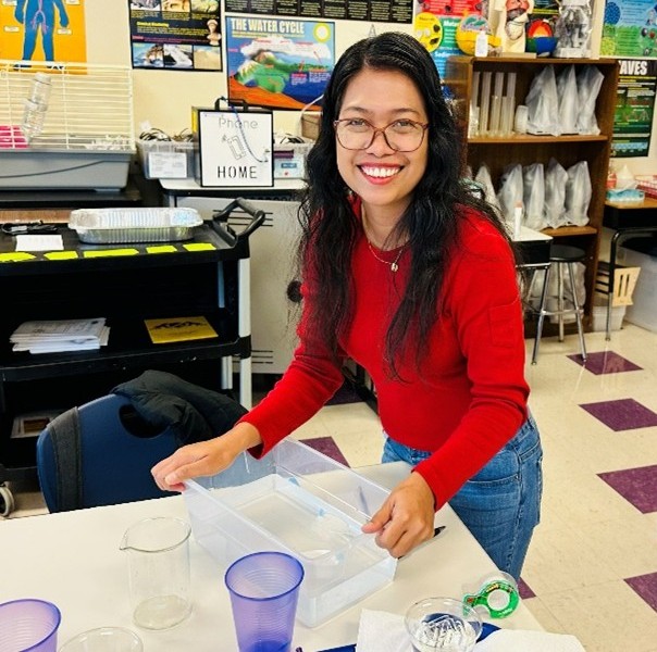 Woman stands by a classroom desk holding equipment for a science experiment, smiling. 